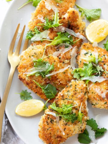 Browned chicken cutlets on a platter, with herb sprigs, shaved Parmesan and lemon wedges.