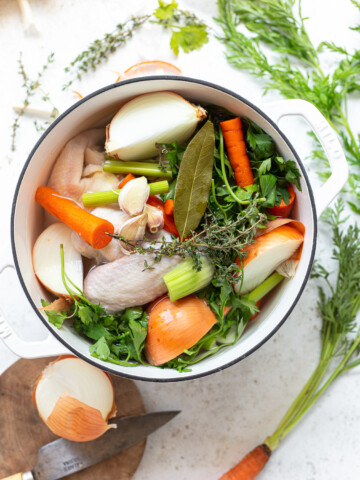 Chunks of onion, carrot, whole chicken wings, bay leaf and herb sprigs in a white pot of water.