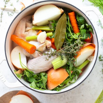 A white pot filled with raw chicken, halved onions, carrots, celery, garlic, parsley, thyme, and a bay leaf—perfect for making rich chicken broth—surrounded by fresh herbs and a partially chopped onion on a cutting board.