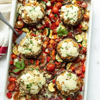 A white baking dish filled with baked stuffed portobello mushrooms topped with melted cheese, surrounded by roasted cherry tomatoes, zucchini, red onions, and red bell peppers—an elegant option for Thanksgiving dinner sides. A serving spoon rests in the dish.