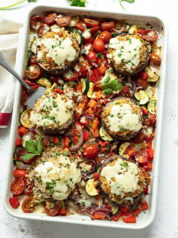 A white baking dish filled with baked stuffed portobello mushrooms topped with melted cheese, surrounded by roasted cherry tomatoes, zucchini, red onions, and red bell peppers—an elegant option for Thanksgiving dinner sides. A serving spoon rests in the dish.