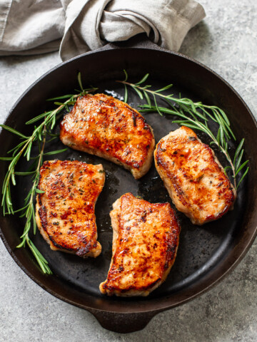 A cast iron skillet with four seared brown sugar pork chops, garnished with fresh rosemary sprigs, rests on a light gray surface next to a folded beige cloth napkin.