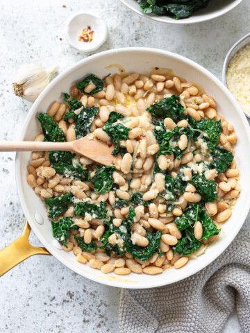 Cannellini beans, kale, and cream sauce in a skillet, with a wood spoon dipping into the pan.