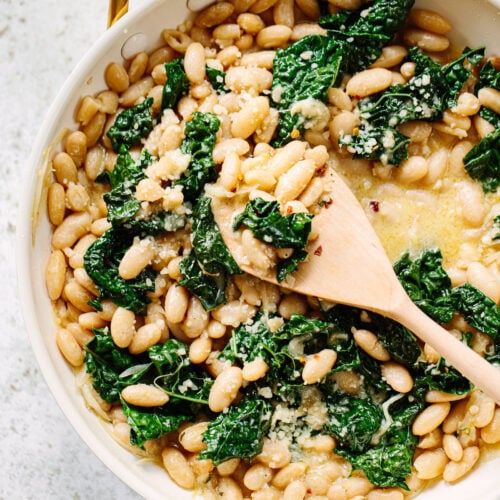 A wooden spoon stirring a pan full of creamy cannellini beans with pieces of wilted kale.