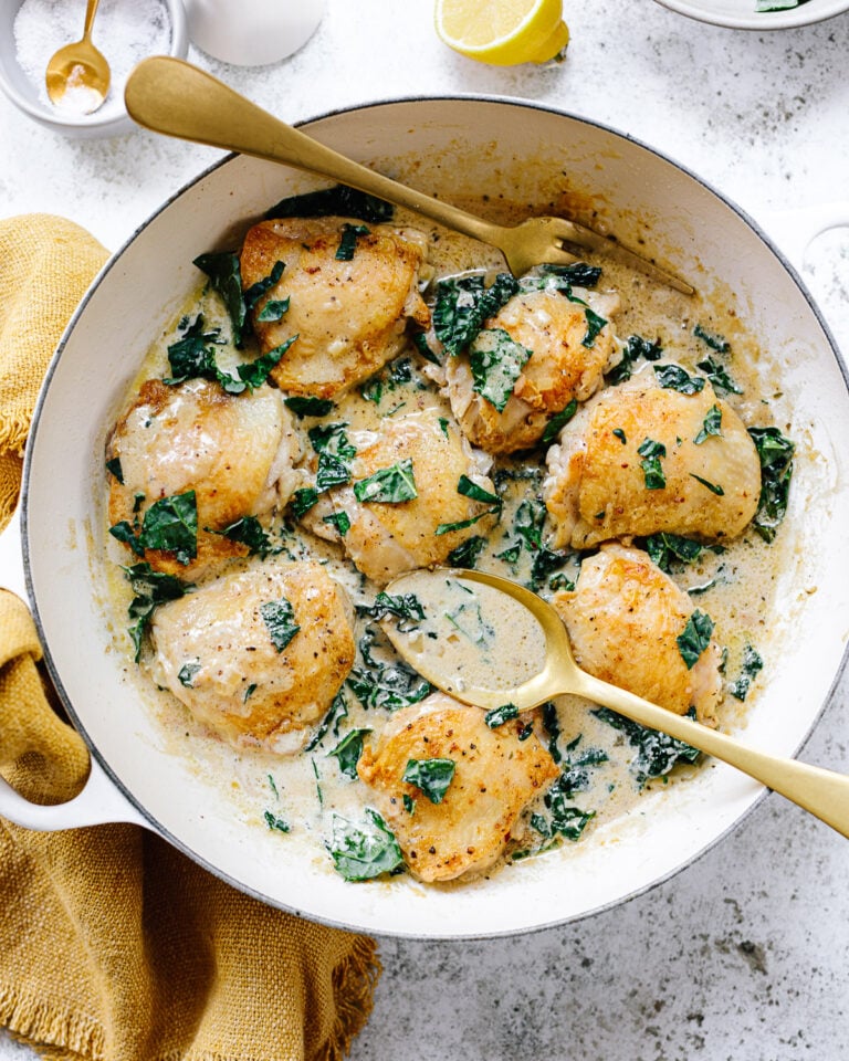 Golden brown chicken thighs in a pan, surrounded by a creamy pan sauce with chopped greens, with a serving fork and spoon alongside.