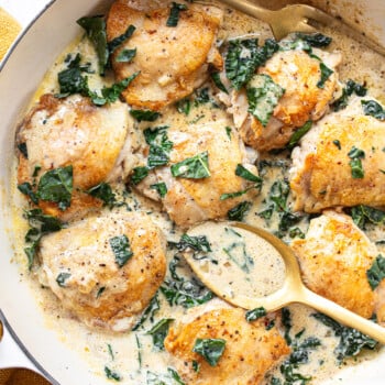 Golden brown chicken thighs in a pan, surrounded by a creamy pan sauce with chopped greens, with a serving fork and spoon alongside.