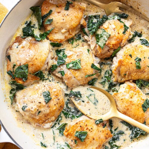Golden brown chicken thighs in a pan, surrounded by a creamy pan sauce with chopped greens, with a serving fork and spoon alongside.