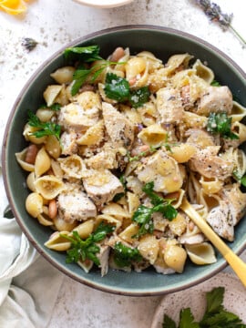 A bowl of pasta shells mixed with chunks of chicken, white beans, fresh parsley, and topped with grated cheese—a twist on classic Italian pasta recipes—served with a gold fork resting in the bowl.