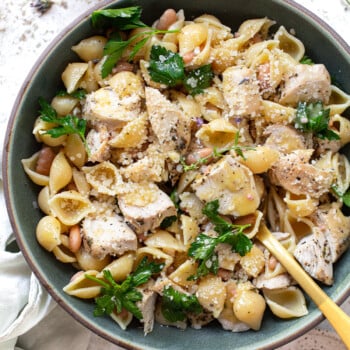 A bowl of pasta shells mixed with chunks of chicken, white beans, fresh parsley, and topped with grated cheese—a twist on classic Italian pasta recipes—served with a gold fork resting in the bowl.