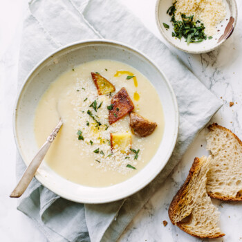 A bowl of creamy potato soup garnished with herbs, croutons, and grated cheese sits on a napkin with a spoon inside. Nearby, a small bowl of herbs and two slices of rustic bread rest on the marble surface.