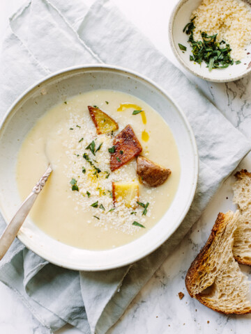 A bowl of creamy potato soup garnished with herbs, croutons, and grated cheese sits on a napkin with a spoon inside. Nearby, a small bowl of herbs and two slices of rustic bread rest on the marble surface.