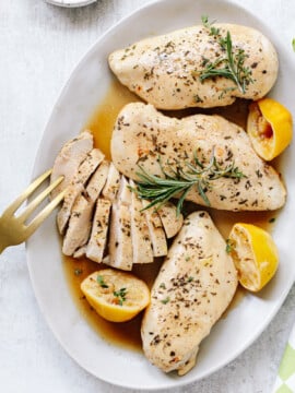 Three whole baked chicken breasts and a sliced chicken breast on a platter, with halved lemons and rosemary sprigs on top.