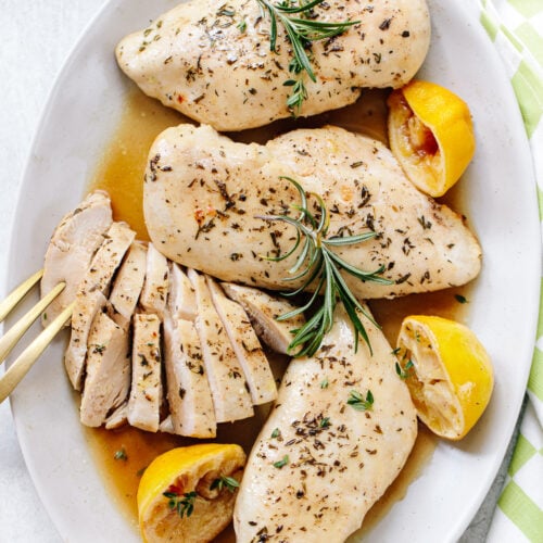 Three whole baked chicken breasts and a sliced chicken breast on a platter, with halved lemons and rosemary sprigs on top.