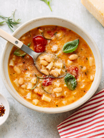 A bowl of creamy bean soup with pasta, white beans, tomatoes, fresh basil leaves, and herbs, served with a spoon. A piece of cheese, a dish of chili flakes, and a red-striped napkin are nearby.
