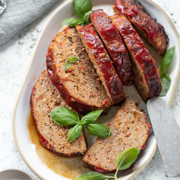 Sliced turkey meatloaf topped with tomato glaze, garnished with fresh basil leaves on a white oval plate. A serving spatula rests beside the slices on a light, textured surface.