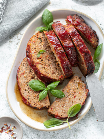 Sliced turkey meatloaf topped with tomato glaze, garnished with fresh basil leaves on a white oval plate. A serving spatula rests beside the slices on a light, textured surface.
