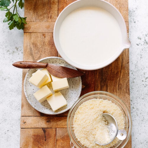 A bowl of heavy cream, sliced butter on a plate, and grated cheese in a cup arranged on a cutting board.
