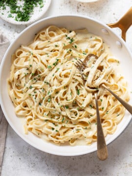 A pan of creamy fettuccine alfredo topped with chopped parsley and grated cheese, with two golden forks twirling the pasta. A bowl of extra chopped parsley sits nearby for the perfect finishing touch.