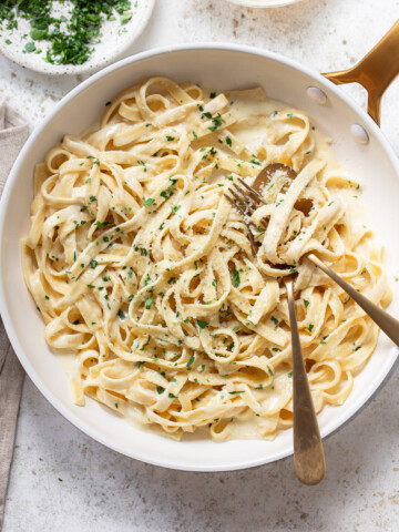 A pan of creamy fettuccine alfredo topped with chopped parsley and grated cheese, with two golden forks twirling the pasta. A bowl of extra chopped parsley sits nearby for the perfect finishing touch.