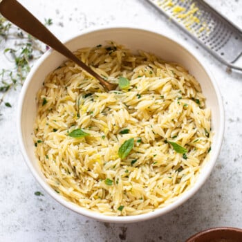A white bowl filled with orzo pasta mixed with herbs and garnished with fresh basil, with a spoon in the bowl. A grater with lemon zest and a small dish of orzo pasta are nearby on a light surface.