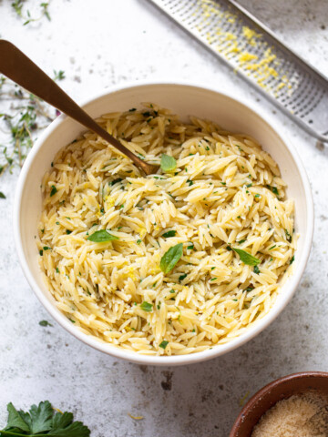 A white bowl filled with orzo pasta mixed with herbs and garnished with fresh basil, with a spoon in the bowl. A grater with lemon zest and a small dish of orzo pasta are nearby on a light surface.