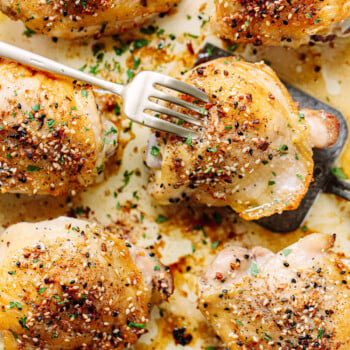 Golden-brown bone-in chicken thighs baked with herbs and spices rest on a baking sheet. A fork pierces one piece, while chopped parsley is sprinkled on top for garnish.
