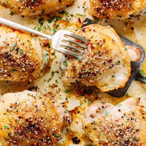Golden-brown bone-in chicken thighs baked with herbs and spices rest on a baking sheet. A fork pierces one piece, while chopped parsley is sprinkled on top for garnish.