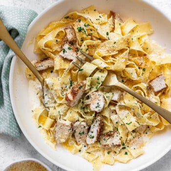 A bowl of creamy chicken alfredo fettuccine pasta topped with sliced grilled chicken and chopped herbs, served with a fork and spoon. Parmesan cheese is in a small bowl nearby on a light-colored surface.