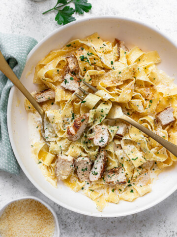 A bowl of creamy chicken alfredo fettuccine pasta topped with sliced grilled chicken and chopped herbs, served with a fork and spoon. Parmesan cheese is in a small bowl nearby on a light-colored surface.