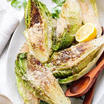 Grilled romaine Caesar salad with dressing and grated cheese on top, arranged on a platter with wood serving spoons, napkins and utensils next to it.