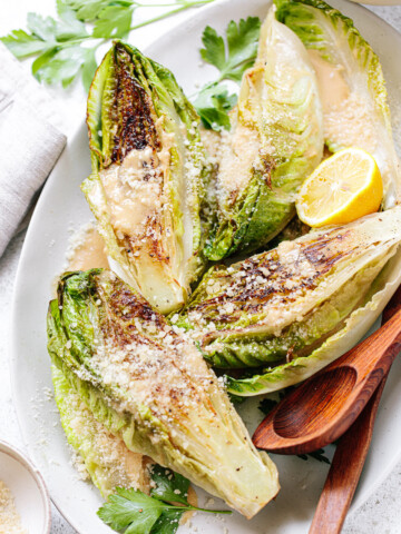 Grilled romaine Caesar salad with dressing and grated cheese on top, arranged on a platter with wood serving spoons, napkins and utensils next to it.