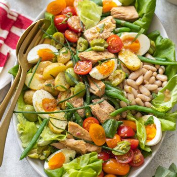 A classic Niçoise salad platter featuring lettuce, cherry tomatoes, green beans, hard-boiled eggs, white beans, olives, and tuna, garnished with herbs and served with salad servers.