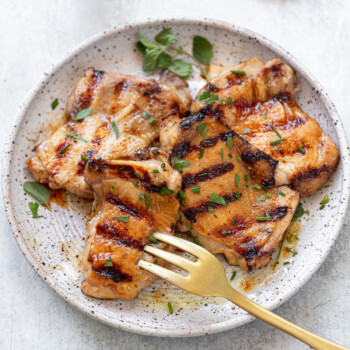 A speckled white plate holds several grilled chicken thighs, infused with a savory chicken thigh marinade and garnished with chopped herbs. A gold fork rests on the plate, and the background includes a light cloth and fresh herb leaves.