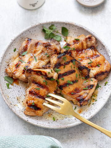 A speckled white plate holds several grilled chicken thighs, infused with a savory chicken thigh marinade and garnished with chopped herbs. A gold fork rests on the plate, and the background includes a light cloth and fresh herb leaves.