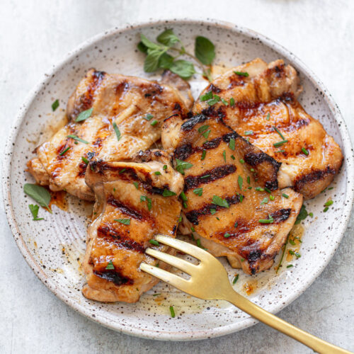 Grilled marinated chicken thighs arranged on a plate, with chopped herbs sprinkled on top and a fork on the side.