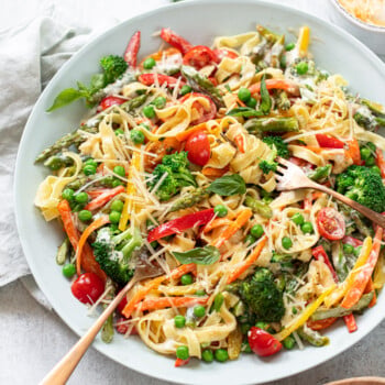 A large plate of vibrant pasta primavera features fettuccine, cherry tomatoes, bell peppers, broccoli, peas, and grated cheese, garnished with fresh basil. Two forks rest on the plate, ready to serve.