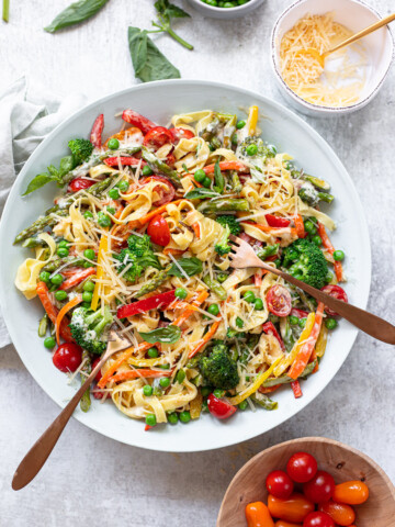 Colorful vegetables on top of pasta in a bowl, with small bowls of cheese, tomatoes and fresh basil sprigs around it.