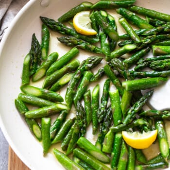 Sautéed asparagus spears in a white skillet with a few lemon wedges, featuring vibrant green colors, sit enticingly on a wooden surface.
