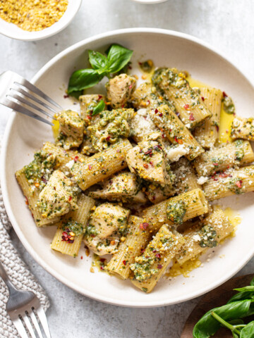 Prepared pasta with pesto chicken sauce in a serving bowl, with sprigs of fresh basil and forks on the side.