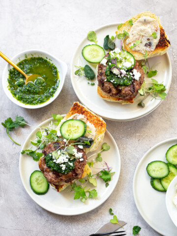 Two burgers arranged open-faced on serving plates, topped with cucumber slices, micro greens, feta cheese and surrounded by bowls of condiments.