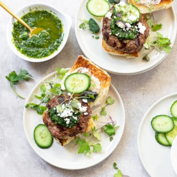 Two burgers arranged open-faced on serving plates, topped with cucumber slices, sprouts, feta cheese and surrounded by bowls of condiments.