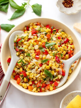 Corn salad with chopped red pepper in fresh basil leaves in a serving bowl, with serving spoons in the bowl.