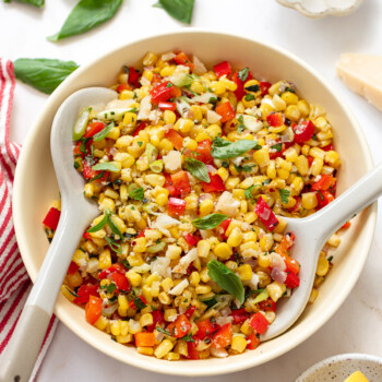 Corn salad with chopped red pepper in fresh basil leaves in a serving bowl, with serving spoons in the bowl.