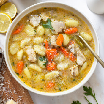 Gnocchi chicken soup in a serving bowl with a spoon, with sliced lemon, hunk of cheese and parsley sprigs in the background.