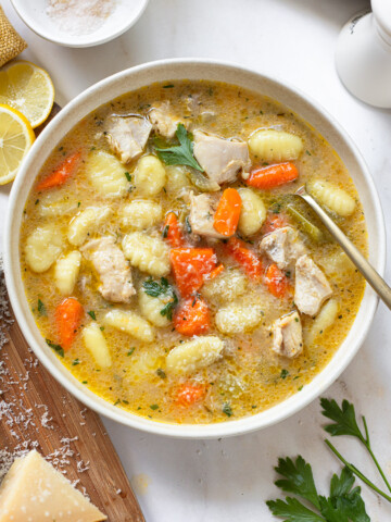 Gnocchi chicken soup in a serving bowl with a spoon, with sliced lemon, hunk of cheese and parsley sprigs in the background.