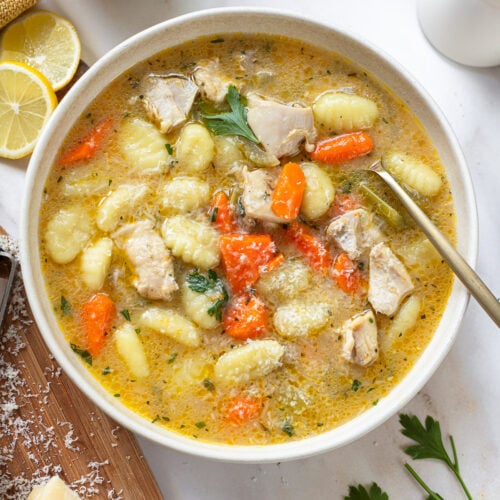 Gnocchi chicken soup in a serving bowl with a spoon, with sliced lemon, hunk of cheese and parsley sprigs in the background.