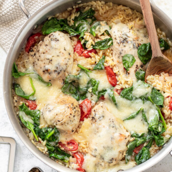 Cooked chicken, orzo, tomatoes and spinach in a skillet, with cherry tomatoes, wooden spoon, spices and a pot holder surrounding it.