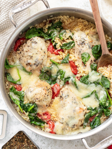 Cooked chicken, orzo, tomatoes and spinach in a skillet, with cherry tomatoes, wooden spoon, spices and a pot holder surrounding it.