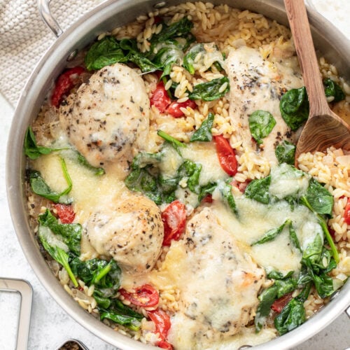 Cooked chicken, orzo, tomatoes and spinach in a skillet, with cherry tomatoes, wooden spoon, spices and a pot holder surrounding it.