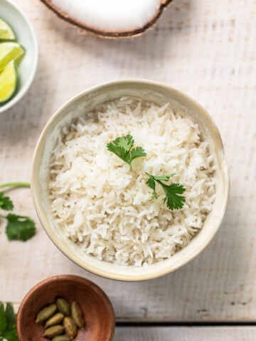 A bowl of cooked coconut rice garnished with fresh cilantro leaves, surrounded by a small bowl of limes, a bowl of cardamom pods, and sprigs of cilantro on a light wooden surface.